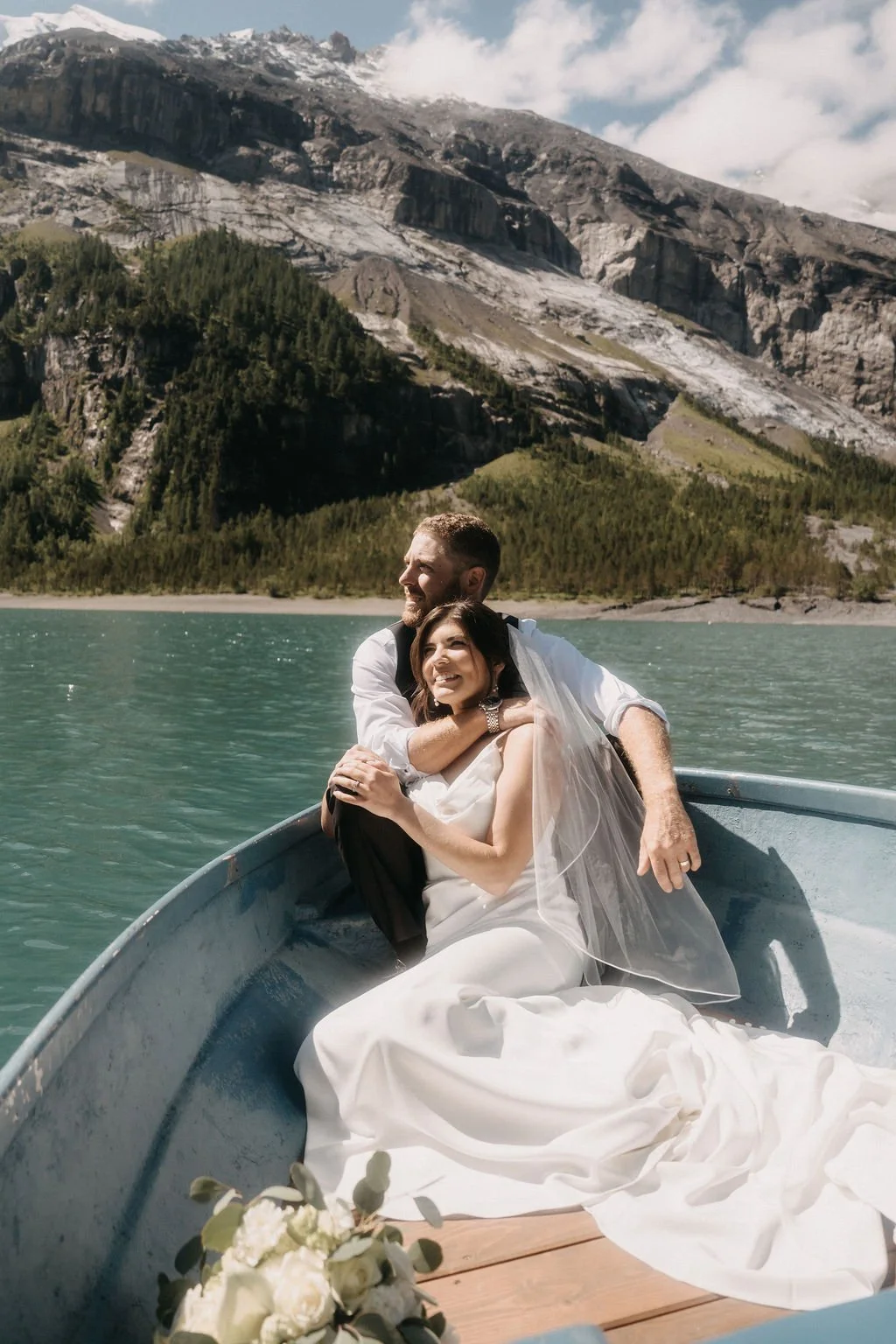 A newlywed couple in wedding attire sitting in a boat on a lake surrounded by mountains and lush green forests.