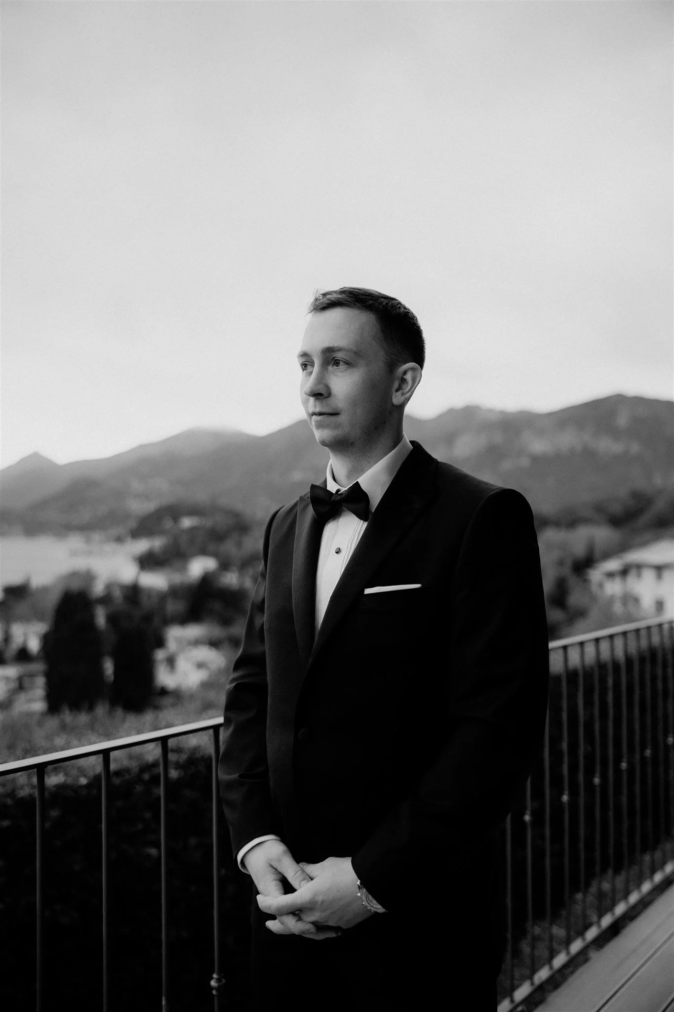 A young man dressed in a tuxedo and bowtie standing outdoors with mountains in the background, viewed in black and white.