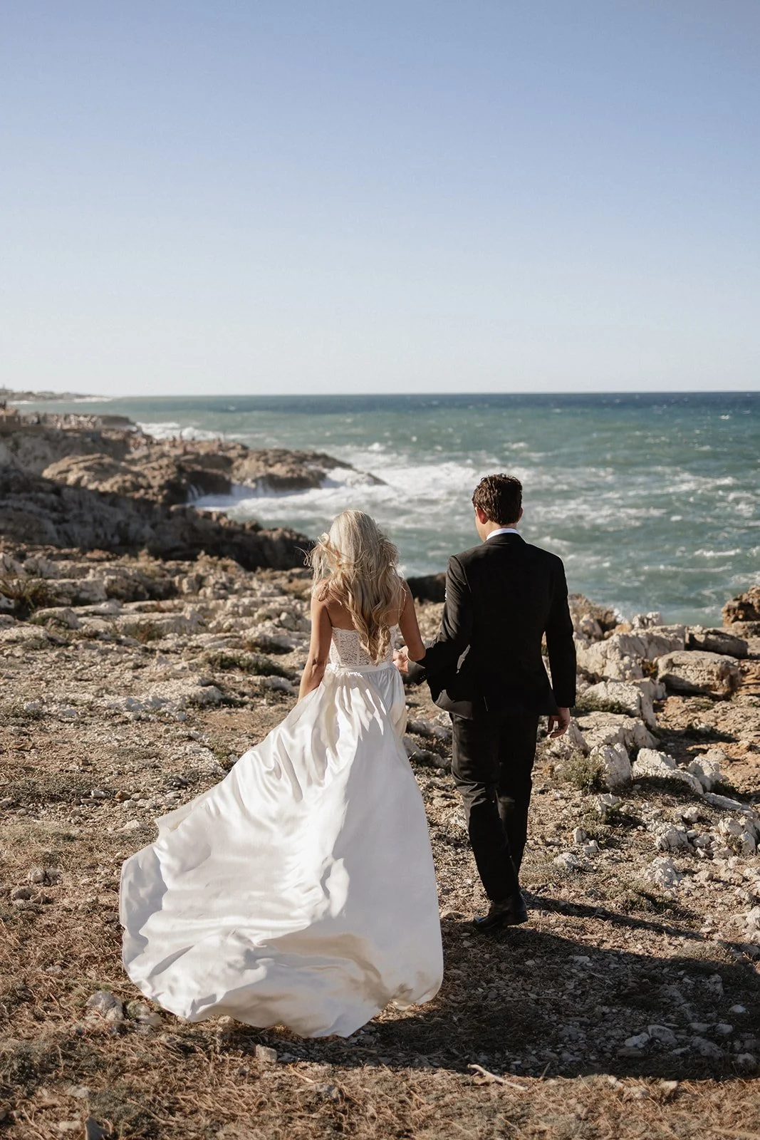 A bride and groom walking hand in hand along a rocky coastline facing the ocean, with clear skies overhead.
