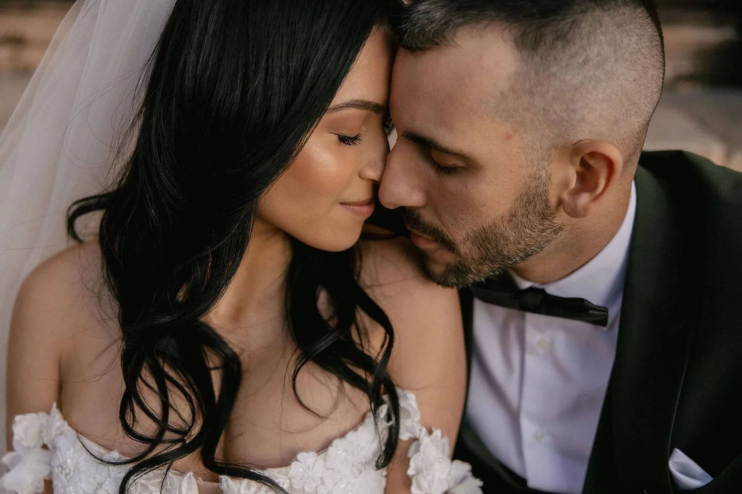 Close-up of a bride and groom with foreheads touching, eyes closed, wearing wedding attire, intimate moment, indoor setting.
