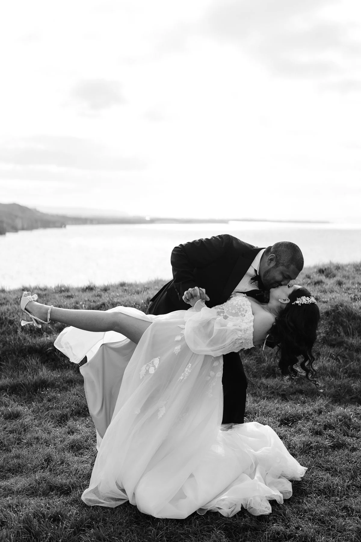 A black and white photograph of a bride and groom outdoors near a body of water. The groom is dipping the bride and kissing her, the bride has her eyes closed and is holding the groom's hand. The bride is wearing a long white wedding dress and a flor