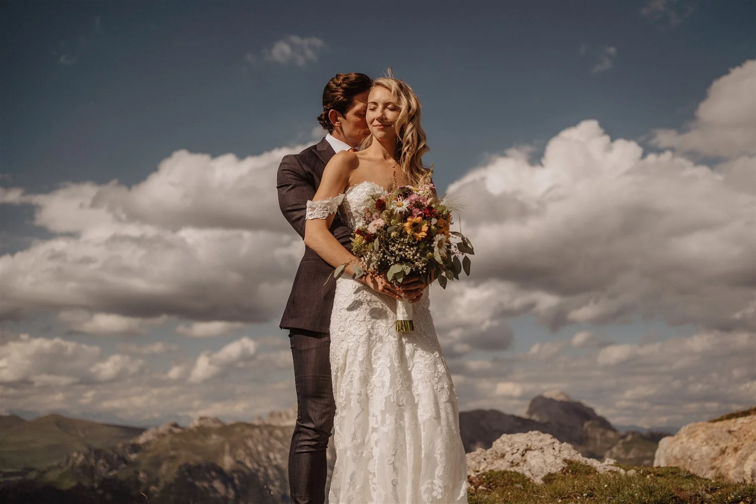A bride and groom standing outdoors in a mountainous landscape, with rocky terrain and cloudy sky. The bride is holding a bouquet of flowers and wearing a white lace wedding dress, while the groom is dressed in a dark suit. The groom is behind the br