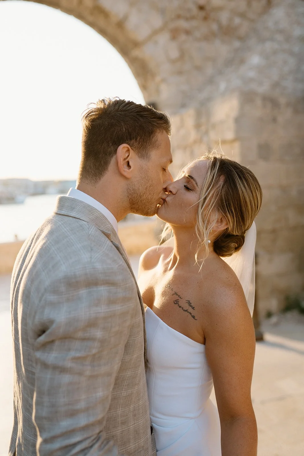 A couple kissing outdoors by a stone archway, with a body of water and city in the background during sunset.