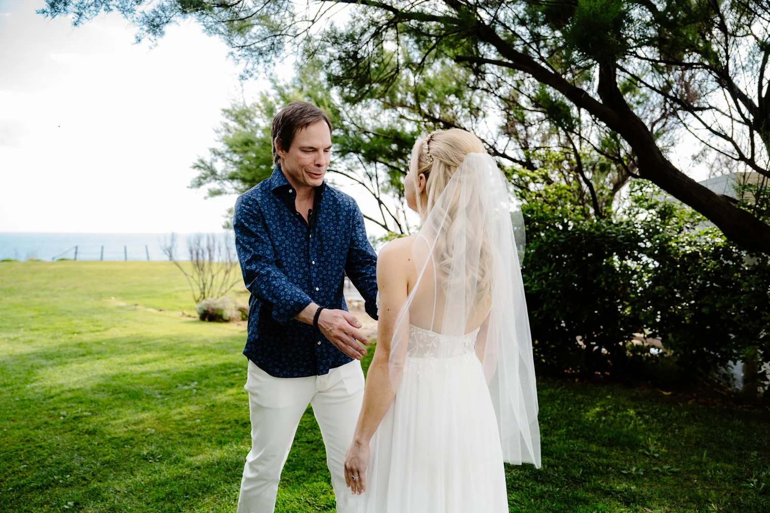 A couple gets married outdoors during the daytime, standing on a grassy area with trees and the ocean in the background. The man is wearing a blue patterned shirt and white pants, holding the woman's hand. The woman is wearing a white wedding dress w