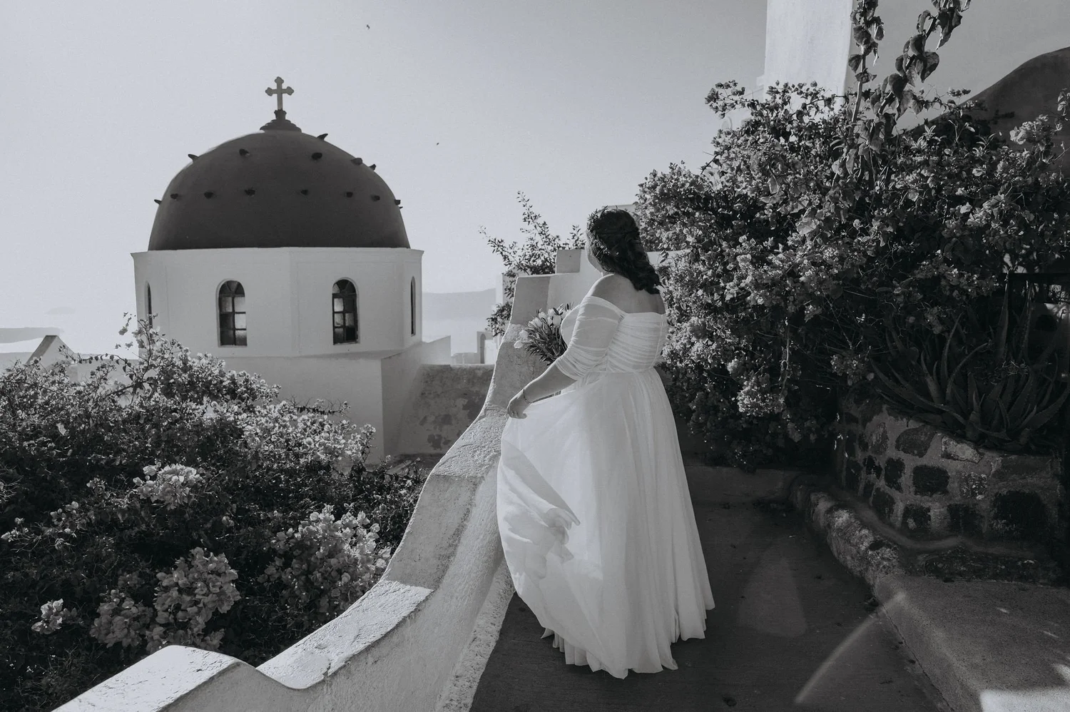A woman in a white off-shoulder dress holding a bouquet standing on a white-walled balcony with a church dome and cross in the background, surrounded by flowering bushes.
