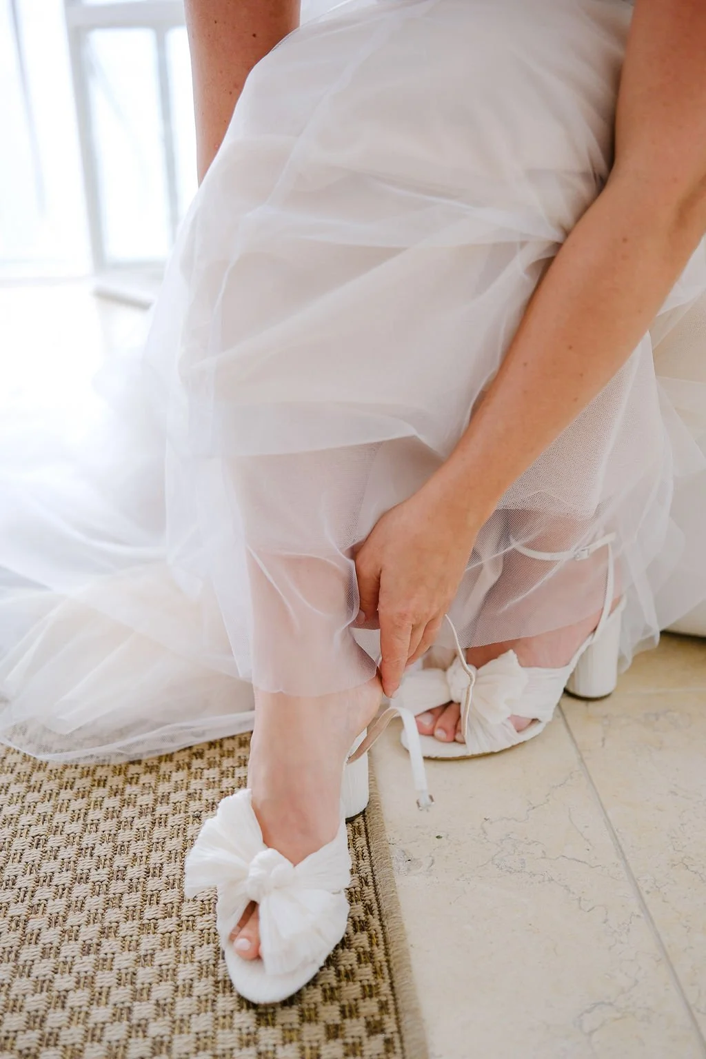 A person in a white dress is adjusting their white high-heeled shoe with a bow on the ankle strap.