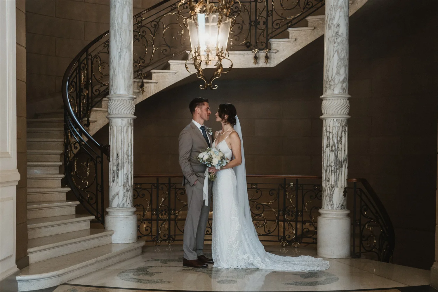 A bride and groom standing close together inside a grand staircase hall, with marble columns, ornate wrought iron railings, and a large chandelier hanging from the ceiling. The bride is dressed in a white wedding gown and holding a bouquet of flowers