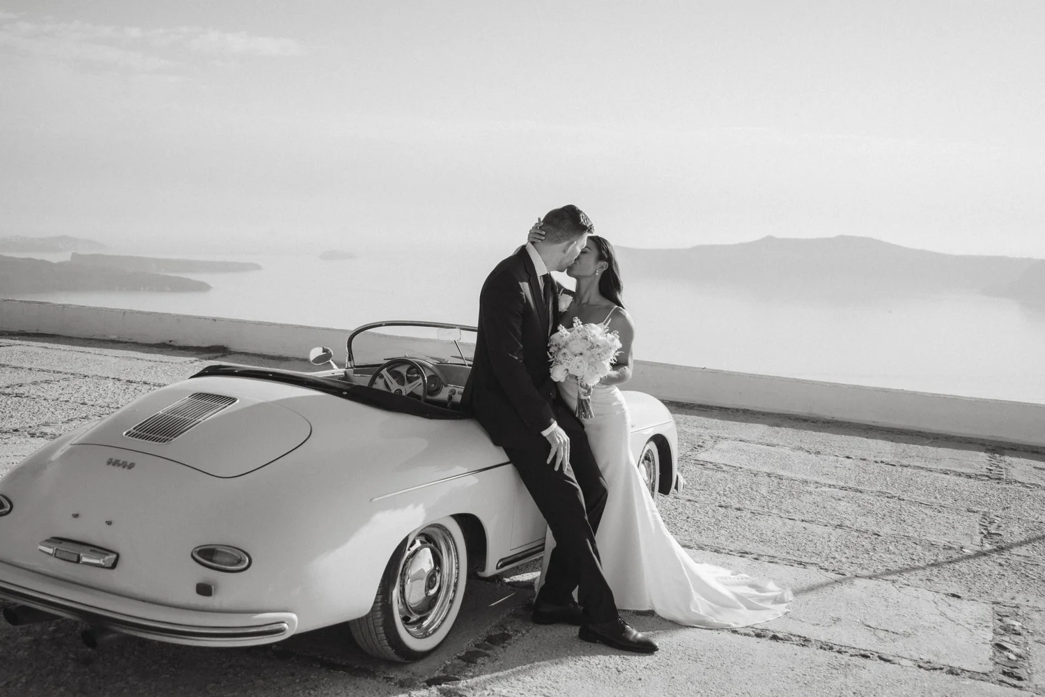 A wedding couple sharing a romantic moment while leaning against a vintage white sports car on a scenic coastal road with mountains in the background.