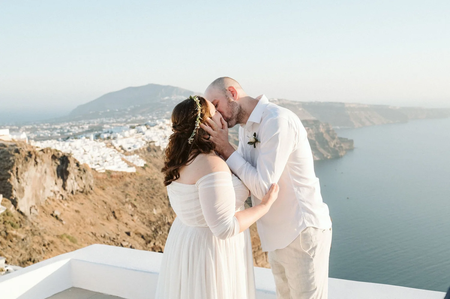 A couple is sharing a kiss on a balcony overlooking the ocean and a coastal town, with mountains in the background. The woman is wearing a white dress and a floral headpiece, and the man is dressed in a white shirt and light-colored pants.