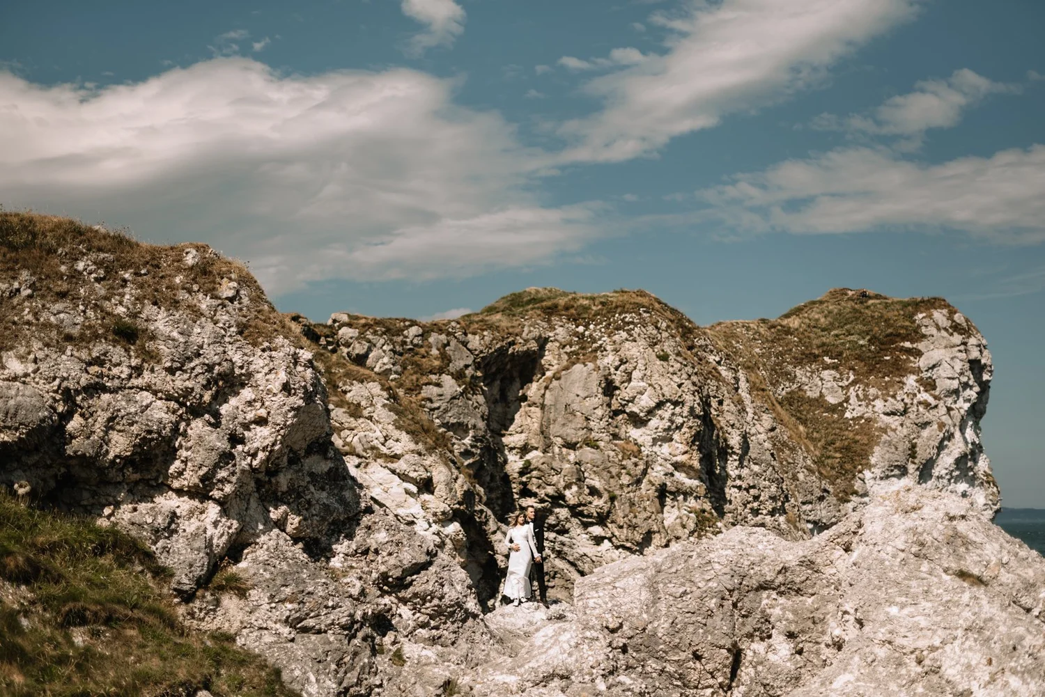 A couple dressed in wedding attire standing on a rocky terrain with cliffs, under a partly cloudy sky.