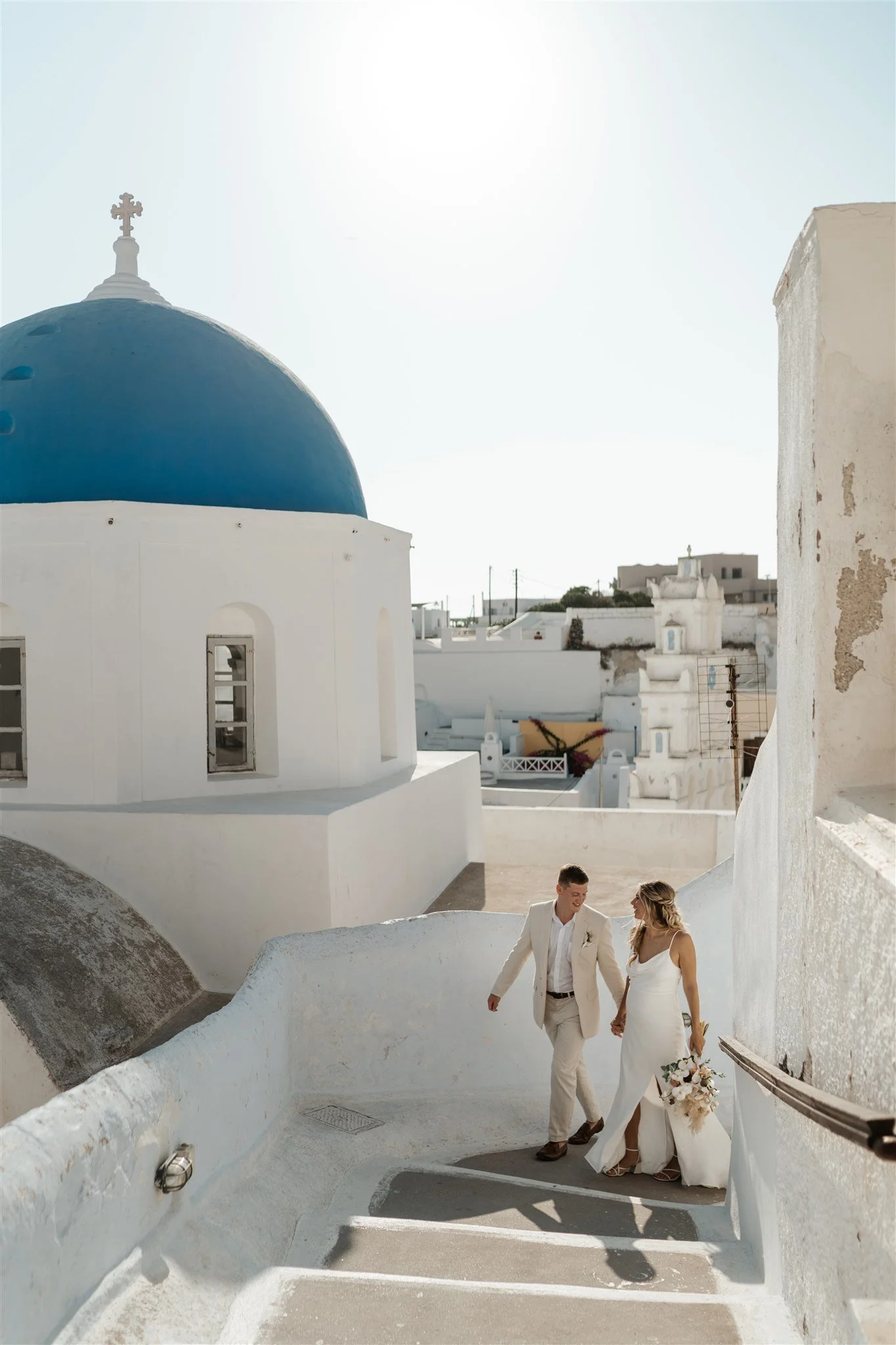 A couple dressed in wedding attire walk hand in hand down a white-washed staircase in Santorini, Greece, with white buildings and a blue-domed church in the background.