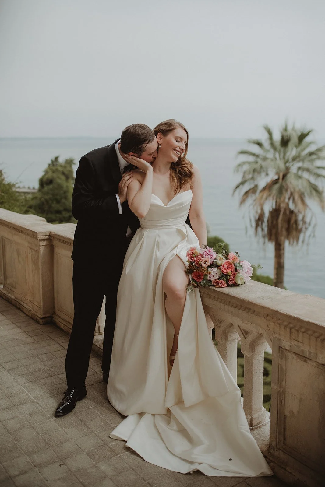 A bride in a white wedding dress with leg showing, holding a pink and white bouquet, and a groom in a black suit with bow tie, embracing and hugging each other on an outdoor balcony overlooking the water and palm trees.