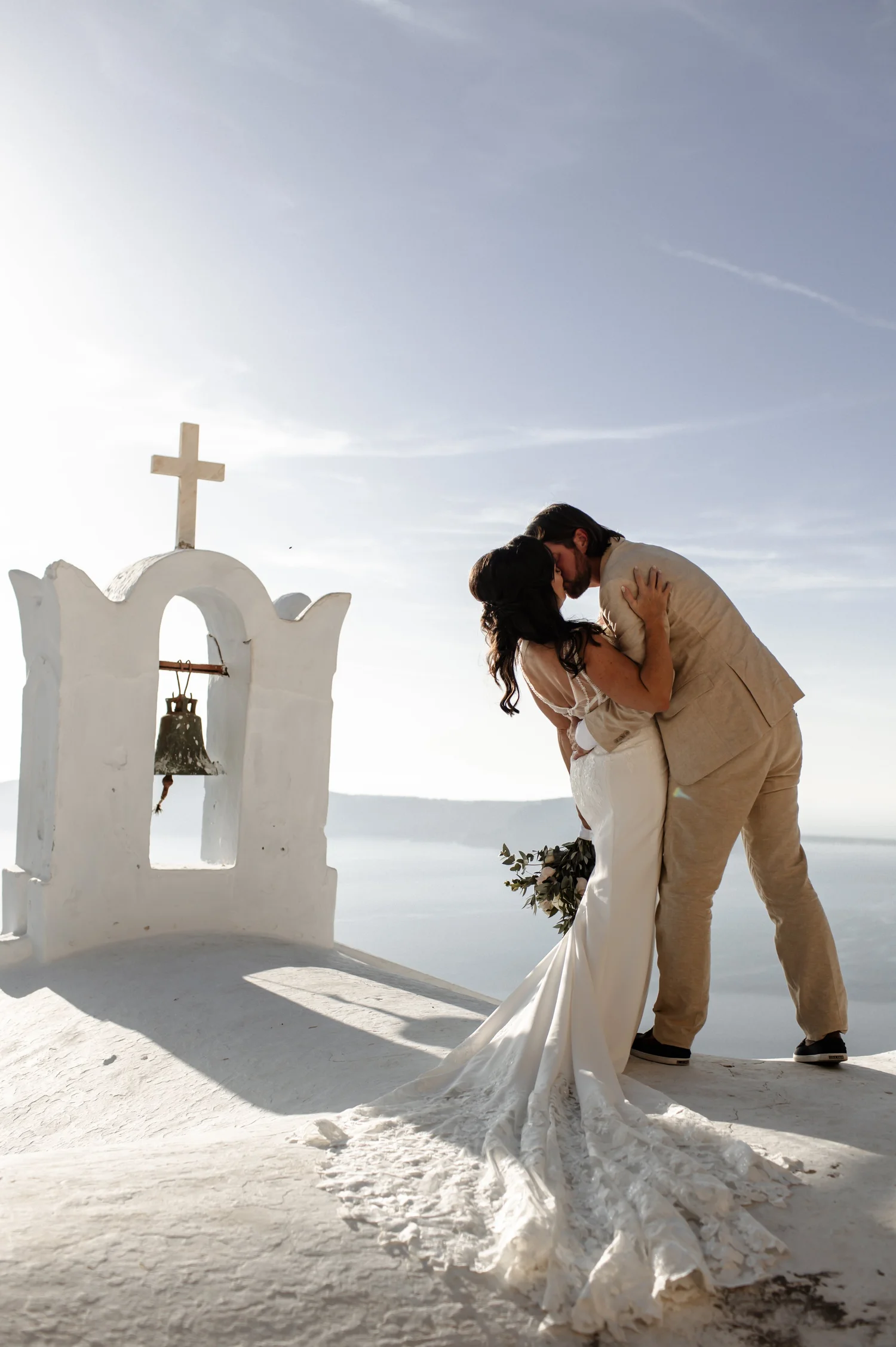A couple sharing a kiss during their wedding in a white dress and beige suit, with a white bell tower with a cross and bell in the background and a clear blue sky.