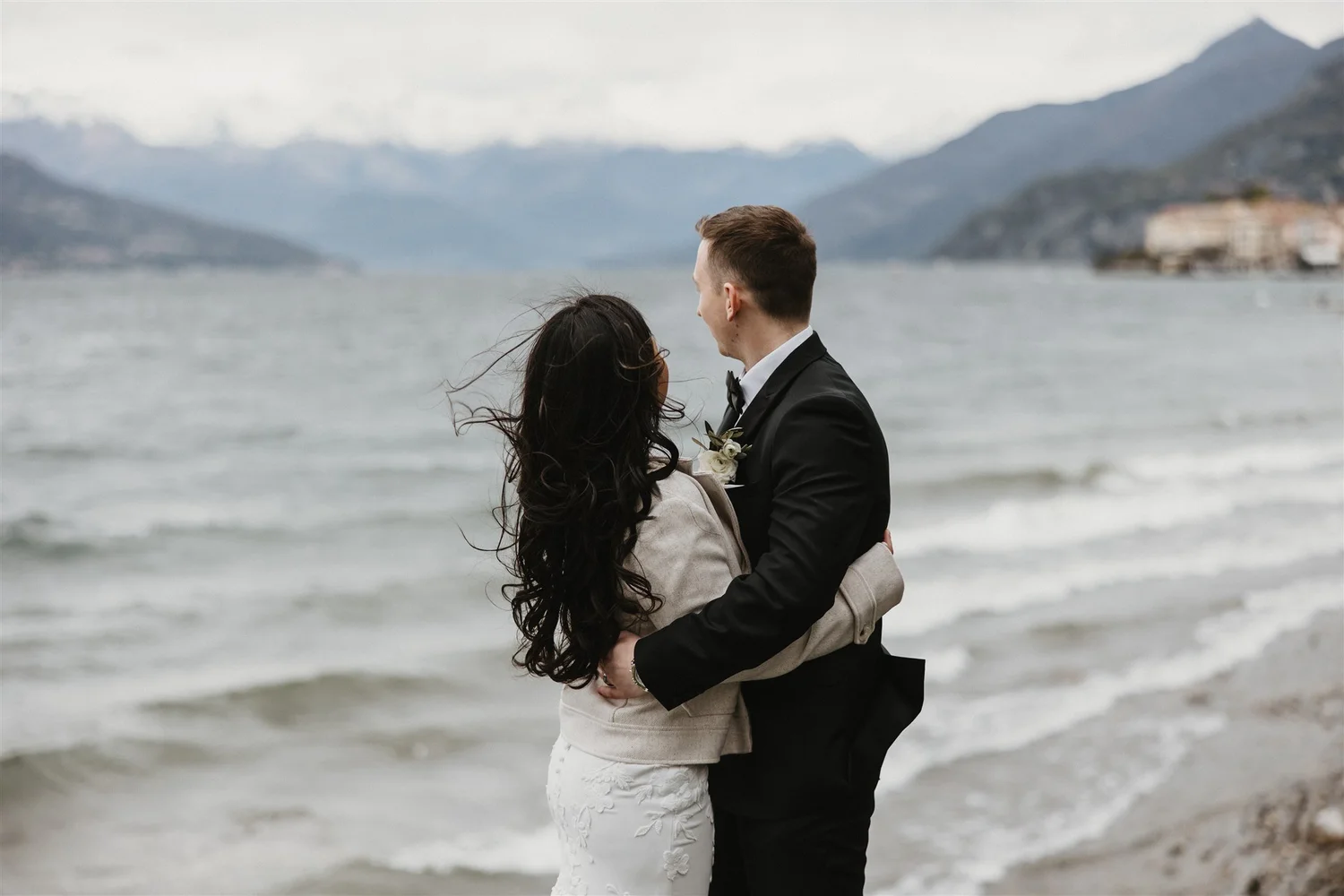 A couple dressed in formal wedding attire embracing on a beach with mountains in the background.