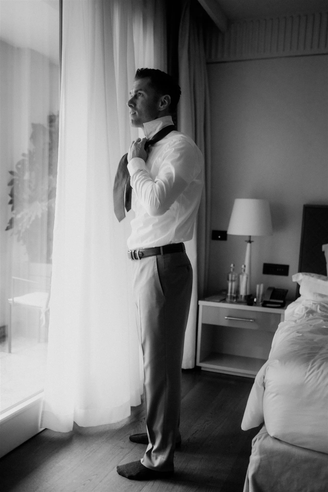 A man in formal attire, adjusting his bow tie, standing by a window with curtains, in a hotel room. He is wearing dress pants, a white shirt, and dark socks. There is a bedside table with water bottles, a lamp, and a television in the background.