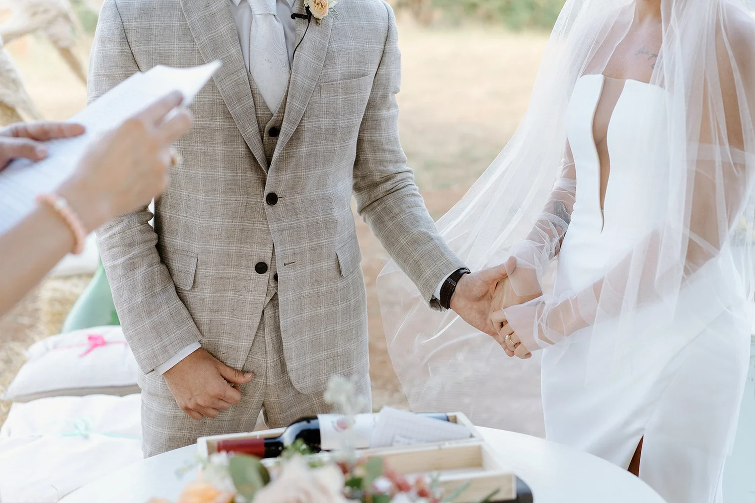 A couple holding hands during a wedding ceremony, with the bride wearing a white dress and veil and the groom in a light-colored suit, outdoors.