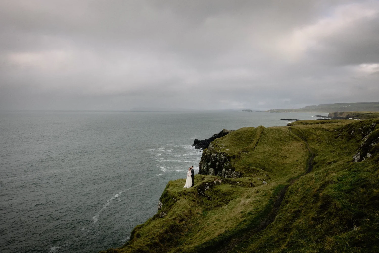 A bride and groom stand on a grassy cliff overlooking the ocean during a cloudy day.