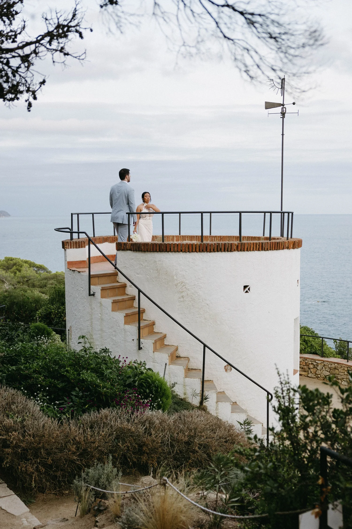A man in a light gray suit and a woman in a white dress stand on a rooftop terrace with a view of the ocean. The woman holds a bouquet and appears to be adjusting her dress. The terrace has a brick parapet and black metal railing, with stairs leading