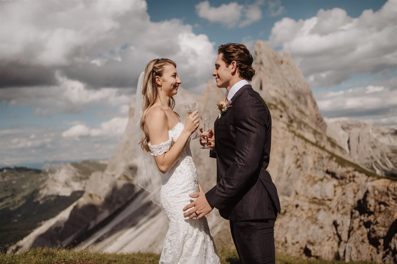 A bride and groom stand face to face, holding wine glasses, with a mountainous landscape and cloudy sky in the background.