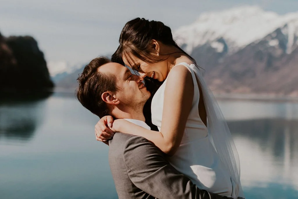 A happy couple embraces near a lake with snow-capped mountains in the background.