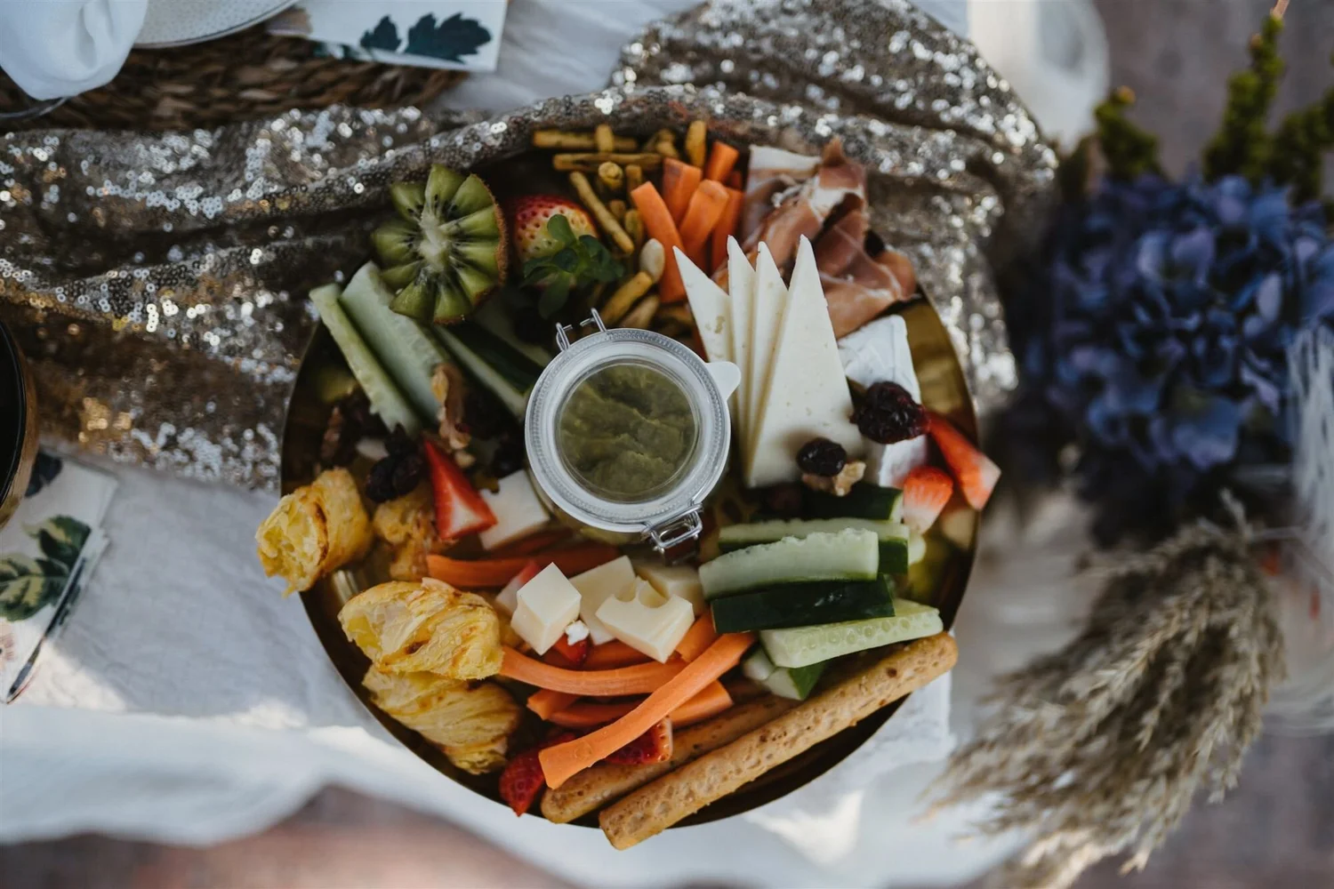 A charcuterie board with assorted cheeses, fresh fruits, vegetables, crackers, and a small container of green dip, partially covered with a silver sequin cloth.