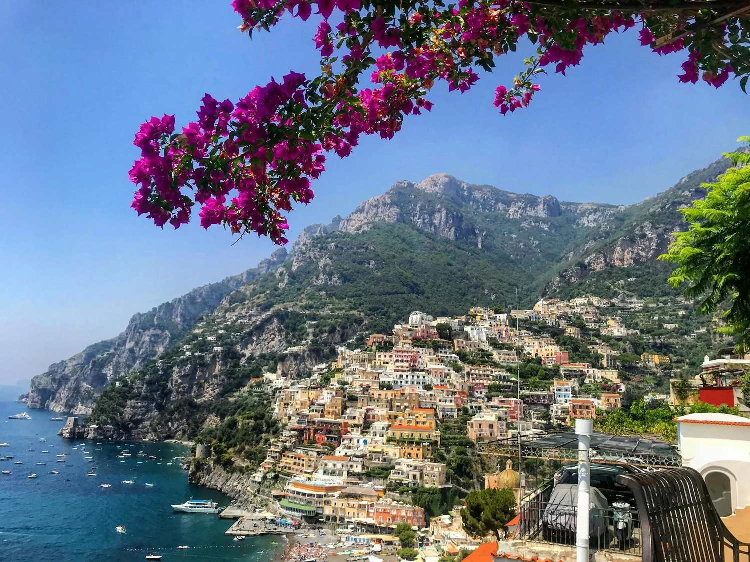 A scenic view of a coastal town built on steep hillside with colorful buildings, mountains in the background, and a harbor with boats, under a clear blue sky. In the foreground, pink flowers and green foliage are visible.