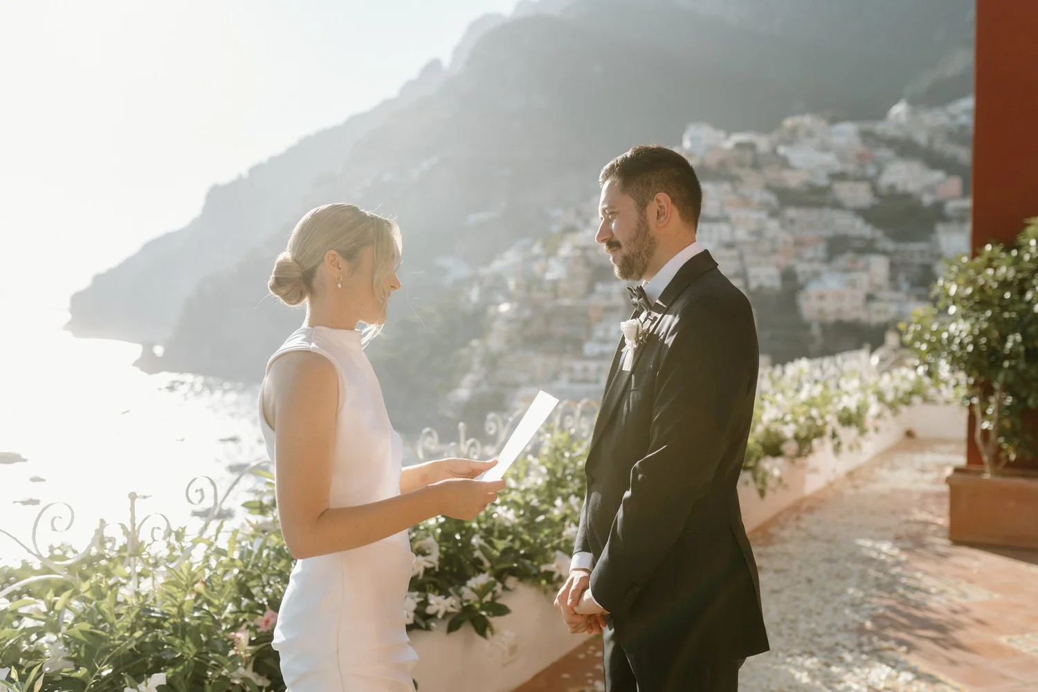A couple stands outdoors during a wedding ceremony, facing each other while holding hands. The woman is wearing a white sleeveless wedding dress, and the man is dressed in a black tuxedo with a bow tie. The background features a scenic hillside with