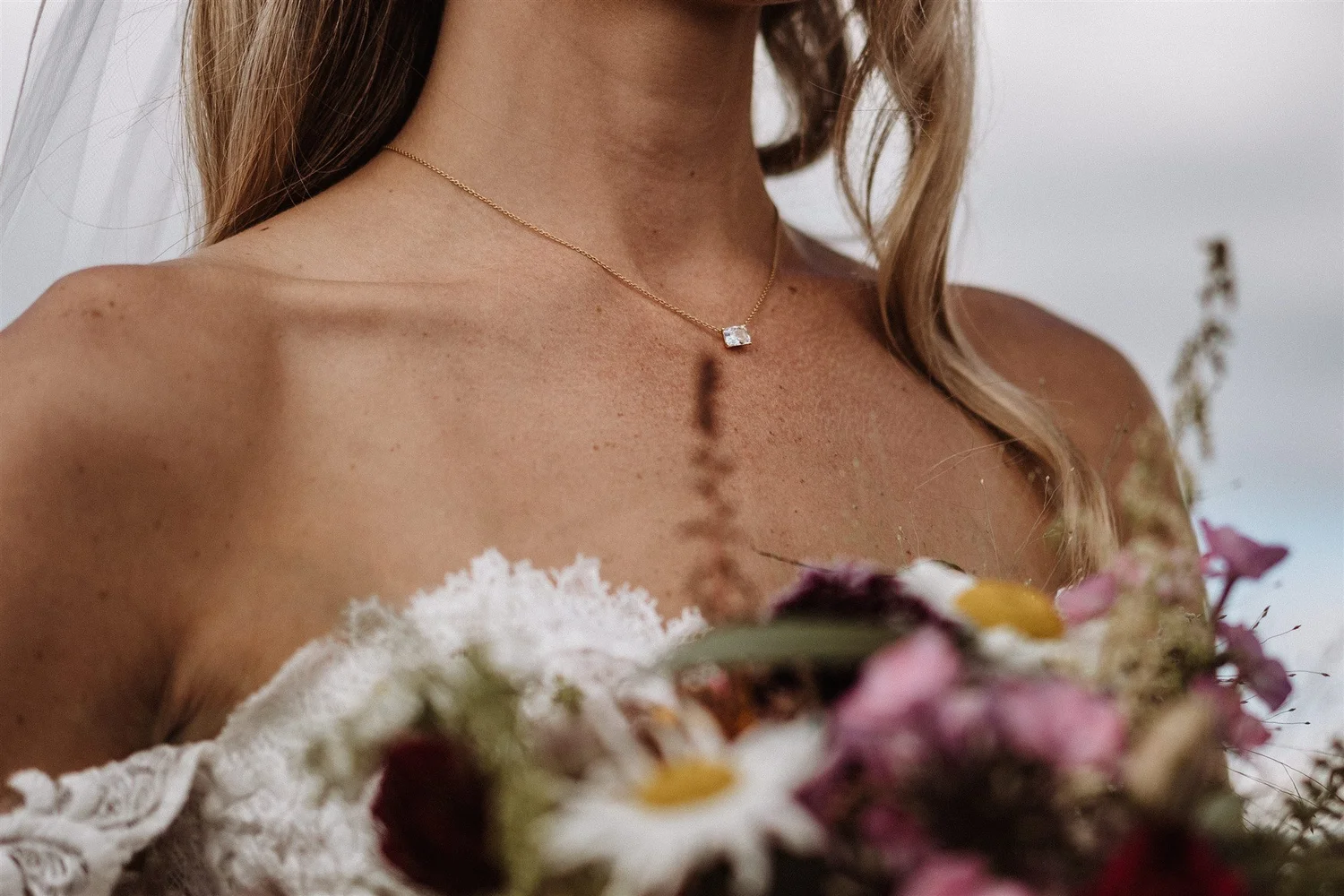 Close-up of a woman wearing a gold necklace with a square-shaped gemstone pendant, holding a bouquet of mixed flowers.