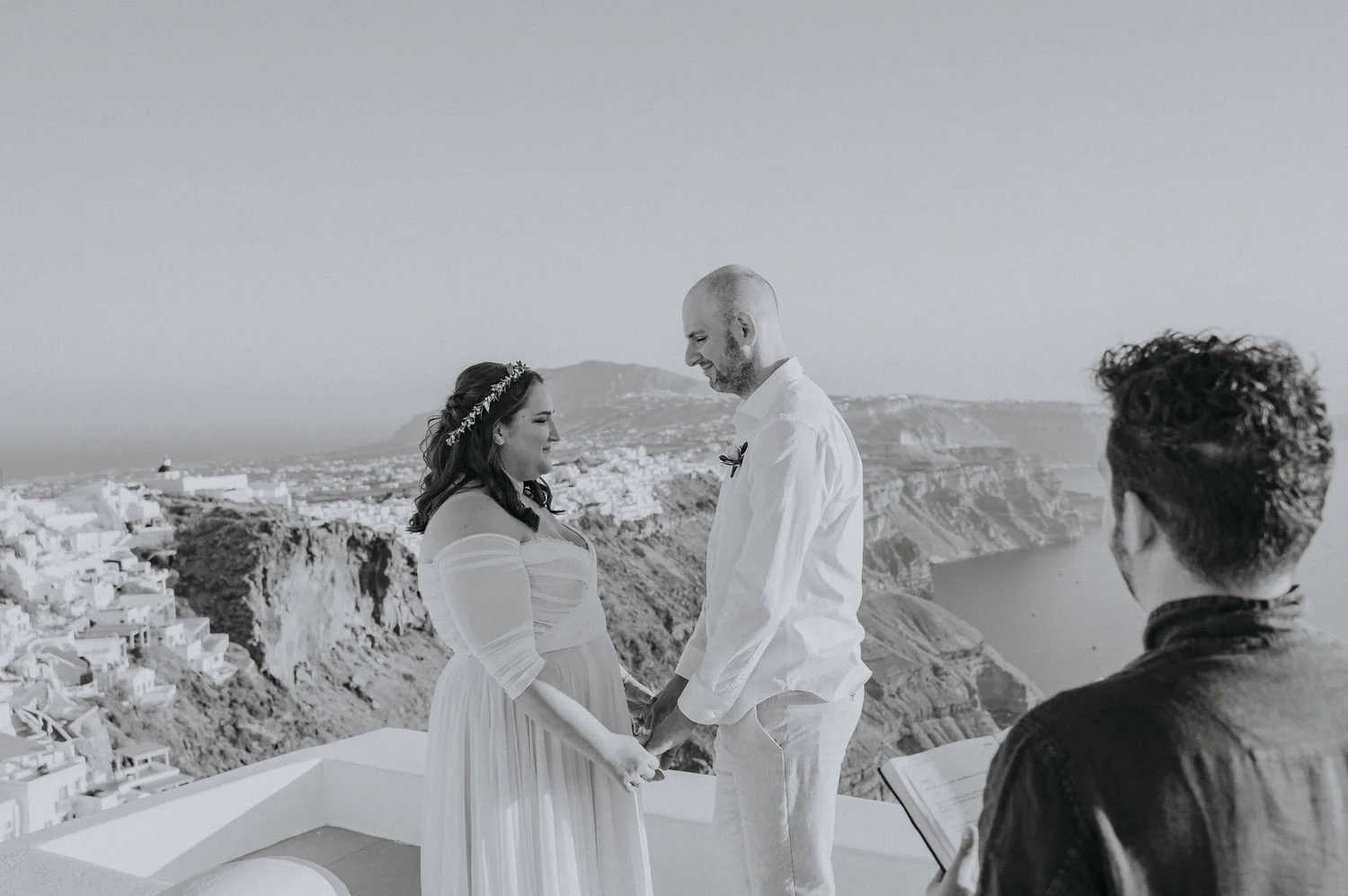 A black-and-white photo of a couple during their wedding ceremony holding hands, with a scenic coastal town and hills in the background, and an officiant on the right.