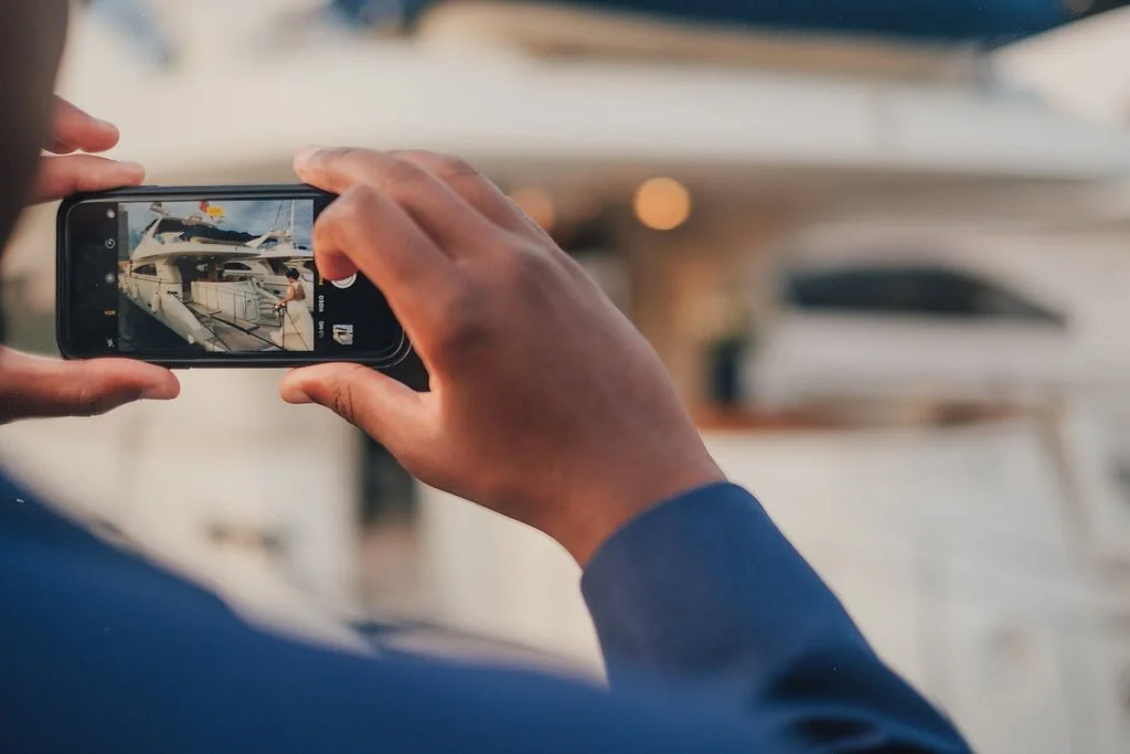 Person taking a photo of a yacht using a smartphone, with a blurred background of boats and water.