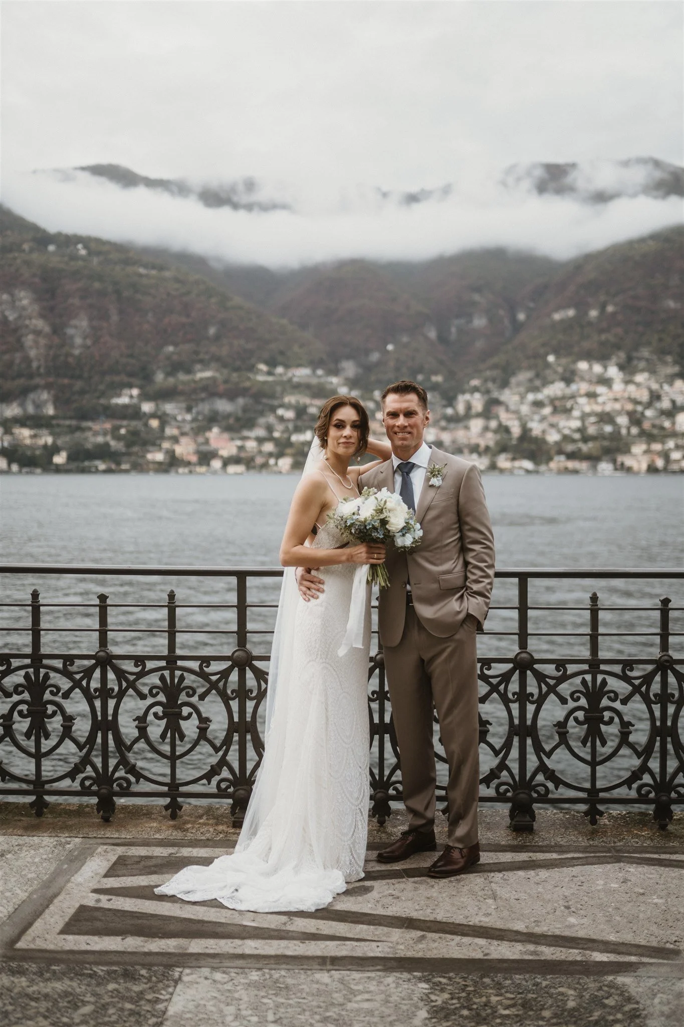 A bride and groom standing together outdoors near a body of water with mountains and houses in the background, dressed in wedding attire, smiling at the camera.