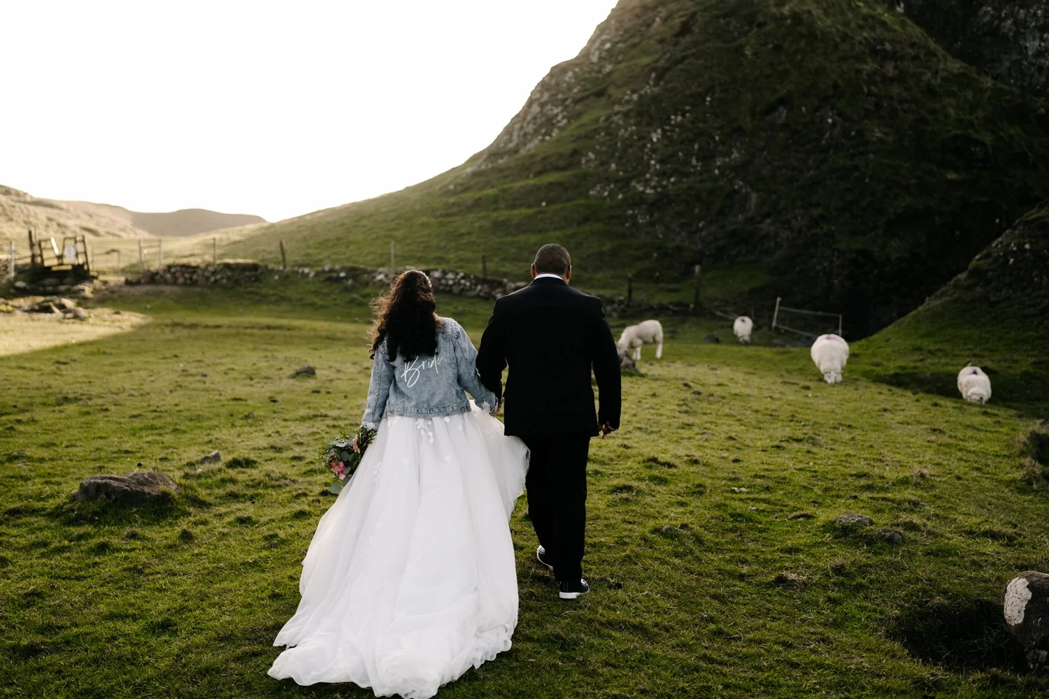 A bride and groom holding hands walking through a grassy field with sheep, surrounded by hills and mountains in the background.