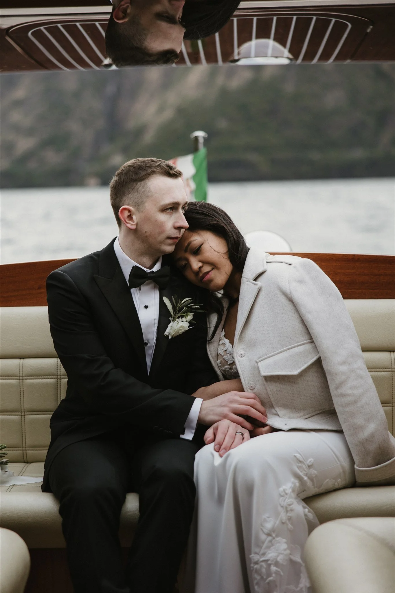 A couple dressed in formal attire sitting close together on a boat, with the woman resting her head on the man's shoulder, and water and a flag in the background.