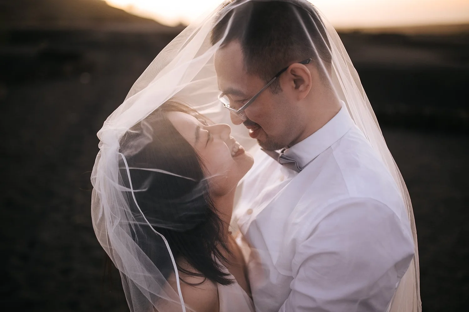 A couple embracing under a wedding veil outdoors at sunset.