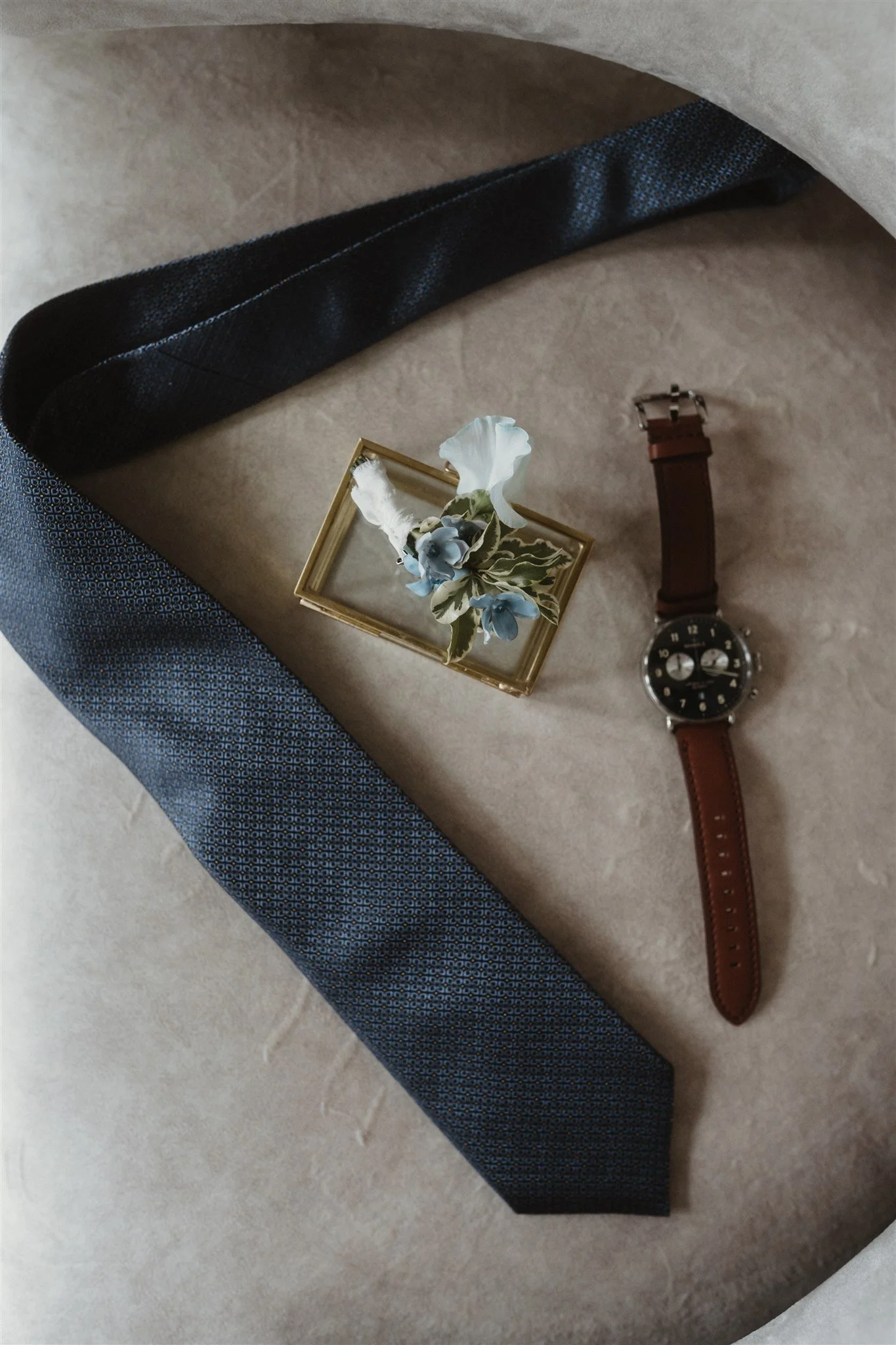 A flat lay of patternless beige fedora hat, dark blue patterned fabric, a brown leather strap watch, gold-framed glass box with blue flowers, and a black belt, arranged on a light-colored surface.