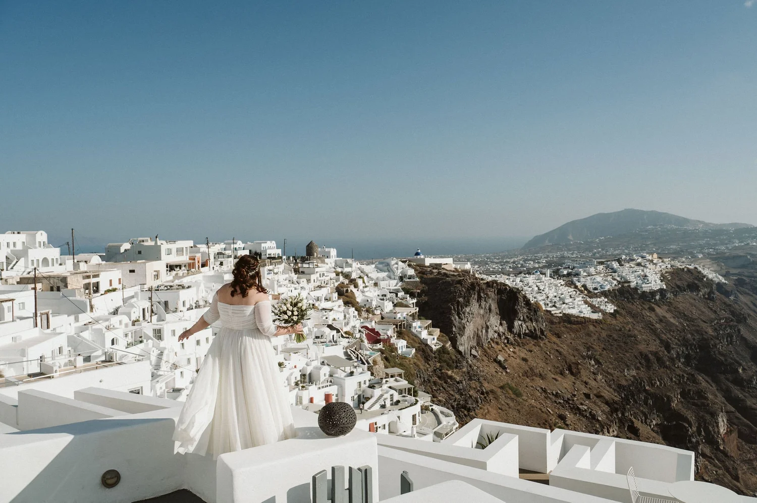 A woman in a white dress holding a bouquet of white flowers standing on a white terrace overlooking a scenic coastal town with white buildings, a small domed church, and a mountain in the background.