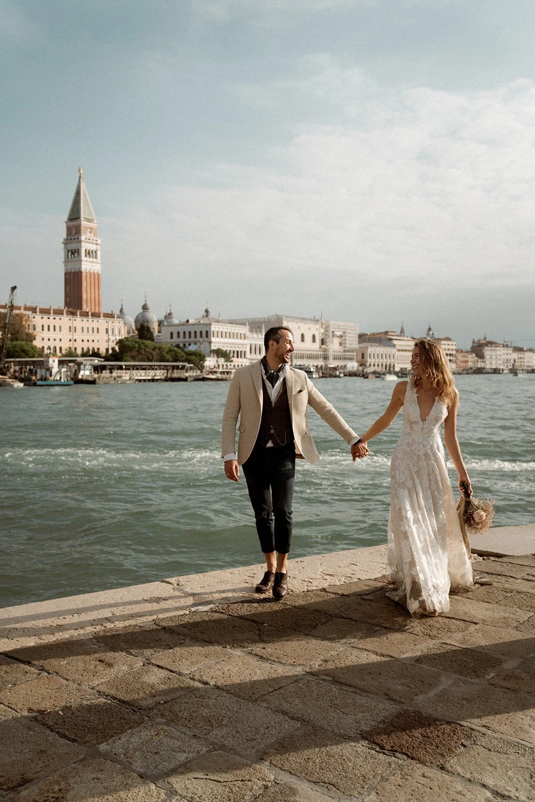 A couple dressed in wedding attire holding hands on a waterfront promenade with a cityscape in the background, including a tall clock tower and historic buildings, under a partly cloudy sky.