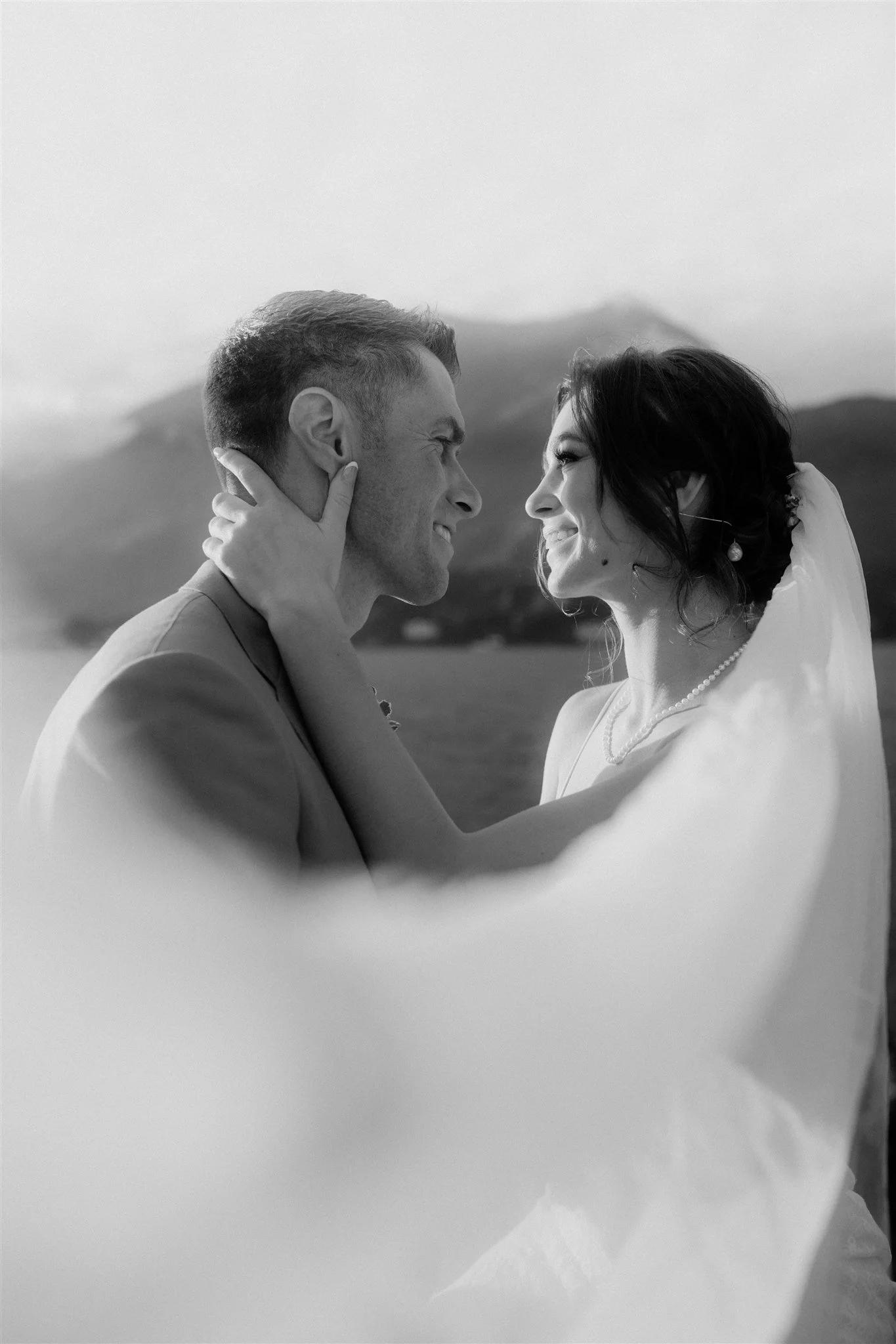 A black and white photo of a bride and groom facing each other, smiling, with the groom holding the bride's face and the bride's hand on the groom's shoulder, outdoors with mountains in the background.
