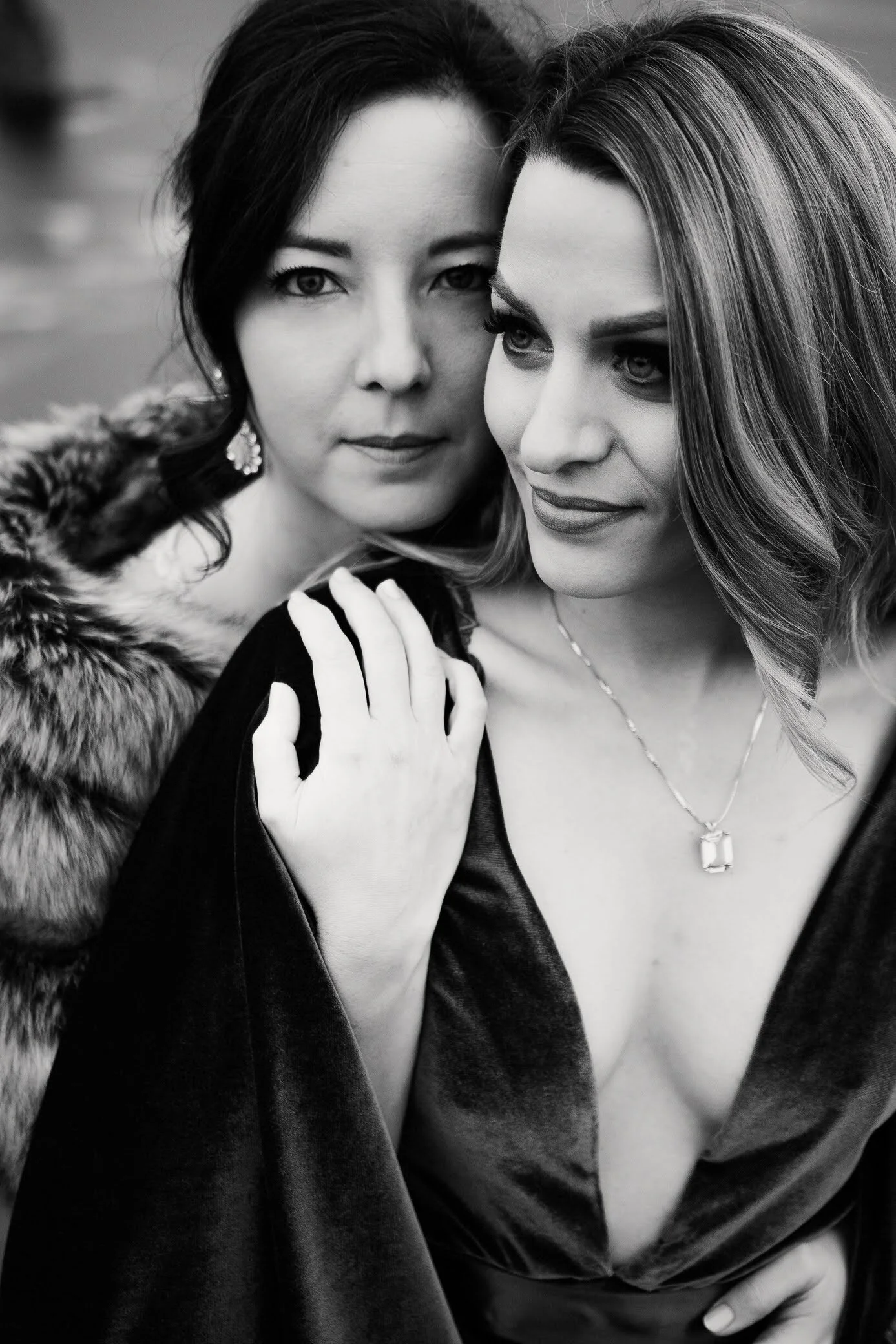 Close-up black and white photo of two women with similar facial features, one wearing a fur coat and jewelry, the other wearing a satin dress with a necklace, standing close together.