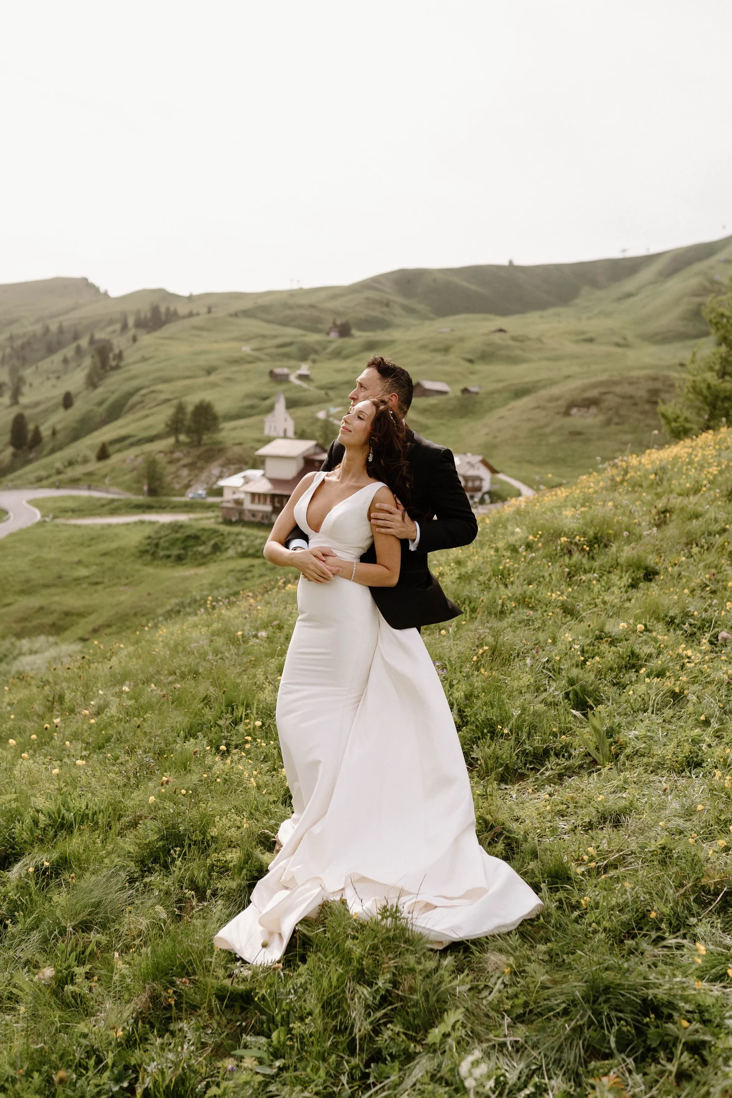 A bride and groom standing together in a green, hilly landscape with small houses and a church in the background. The bride is wearing a white wedding dress, and the groom is in a dark suit. They are embracing each other with scenic hills behind them