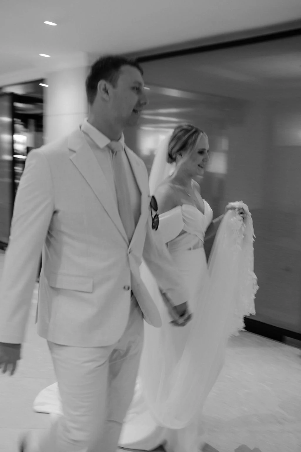 A black and white photo of a bride and groom walking in an indoor setting, both dressed in wedding attire.