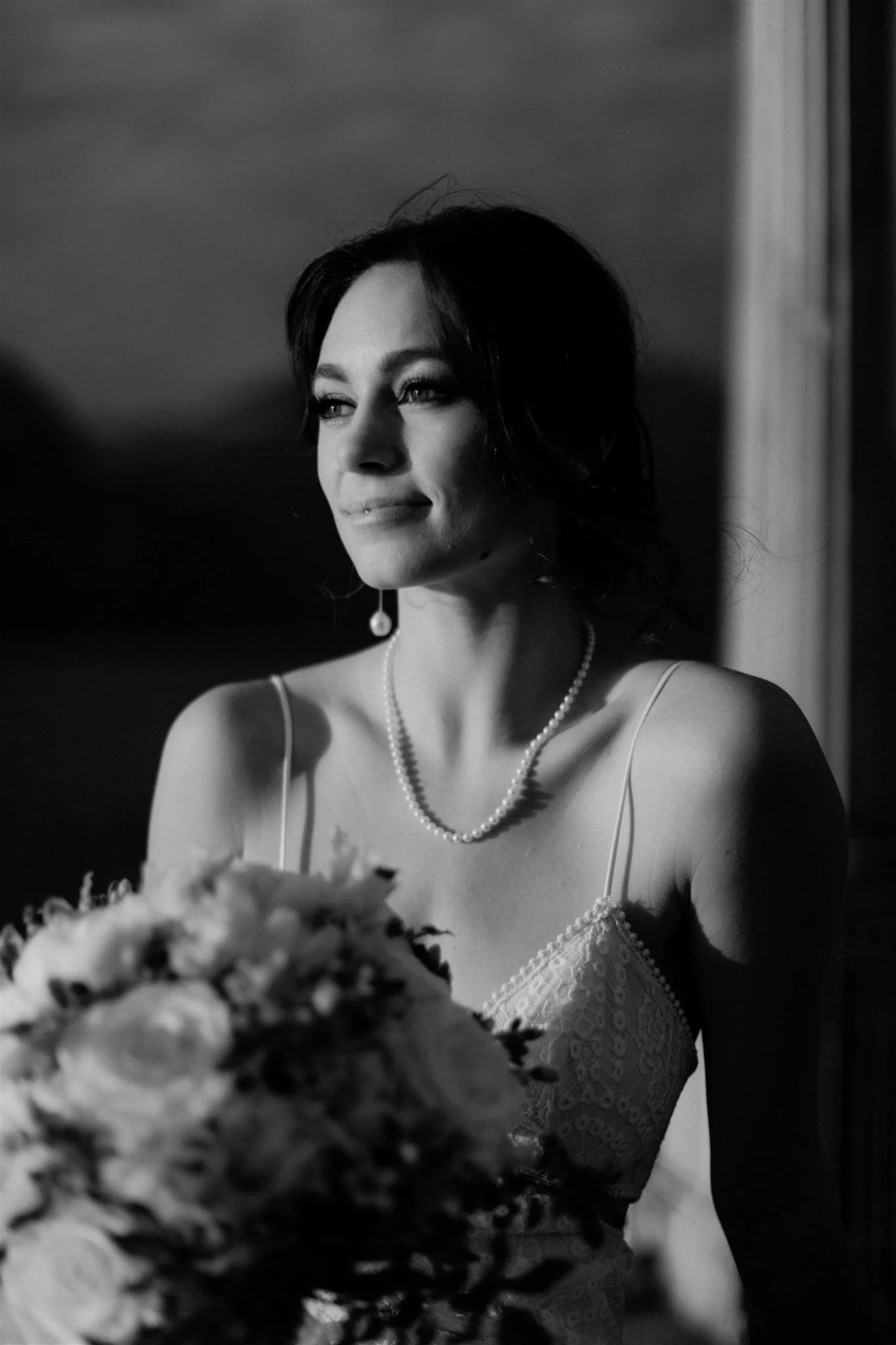 A black-and-white photo of a woman wearing a white lace dress, a pearl necklace, and a pearl earring, smiling softly while looking to the side. She is positioned near a window with light coming through.