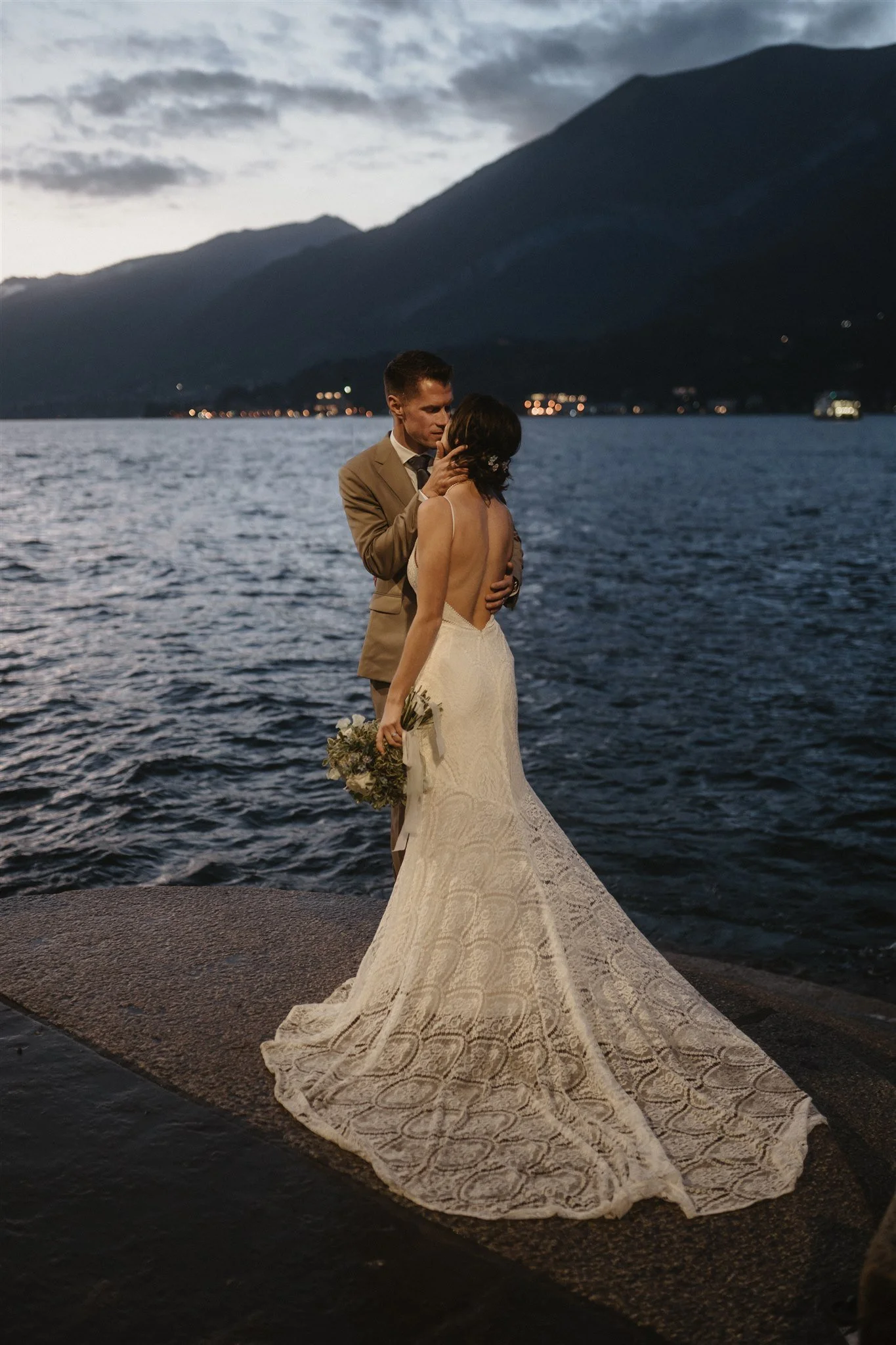 A bride and groom embrace near a body of water at dusk, with mountains and a cloudy sky in the background. The bride wears a lace wedding gown and holds a bouquet, while the groom wears a tan suit.