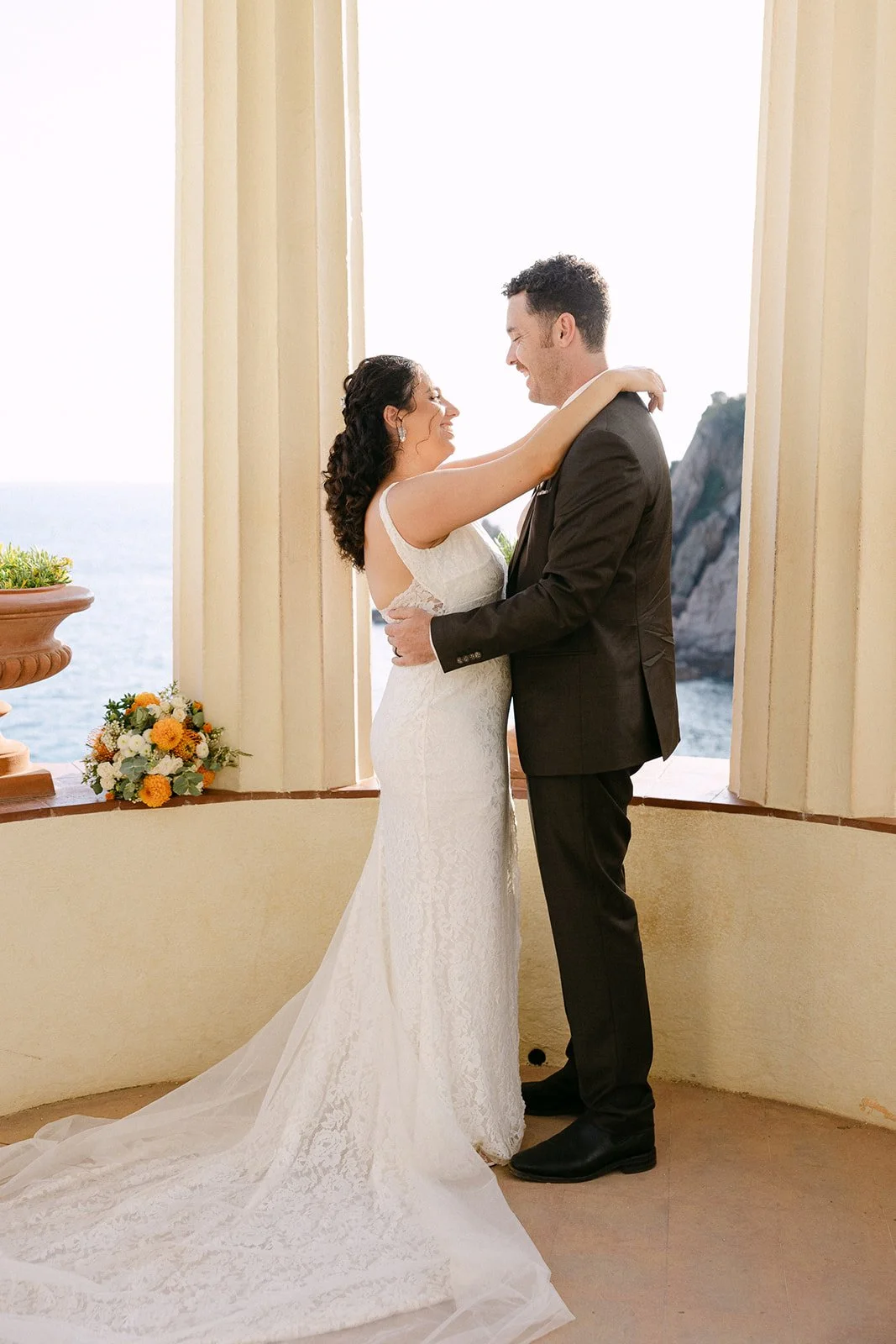 A bride and groom smiling and looking at each other, standing close in a bright room with large windows and a scenic ocean view.