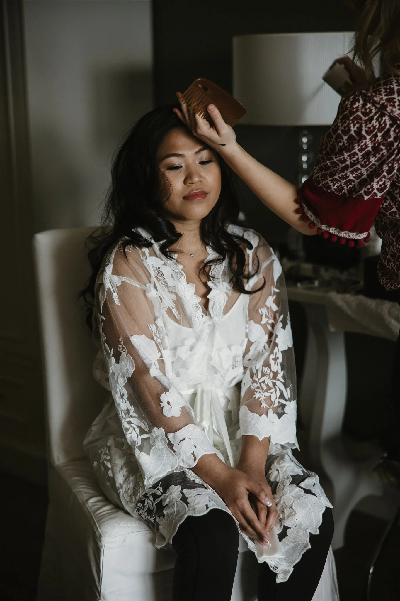 A woman with dark hair is sitting with her eyes closed, wearing a white lace robe. Someone is gently combing her hair with a brown comb.