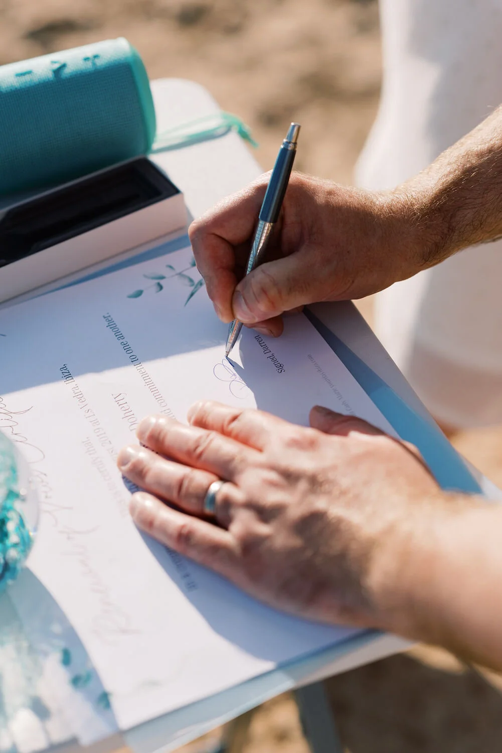 A person is signing a document with a pen on a table outdoors, with a wedding cake and a turquoise speaker in the background.
