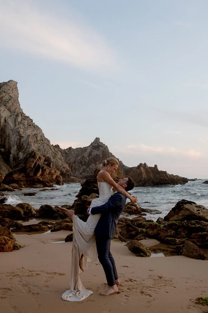 A couple on a beach, with the man lifting the woman in his arms as they look at each other, surrounded by rocks and cliffs.