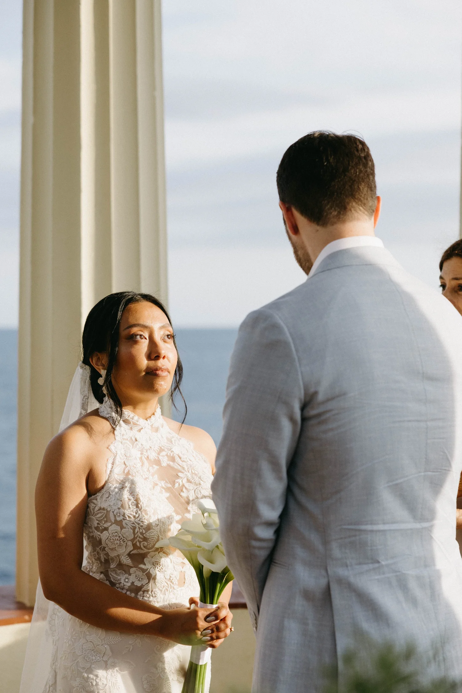 A woman in a wedding dress holding a bouquet of white calla lilies, standing at an outdoor wedding ceremony, with a man in a light gray suit facing her, and a partially visible woman with dark hair on the right.