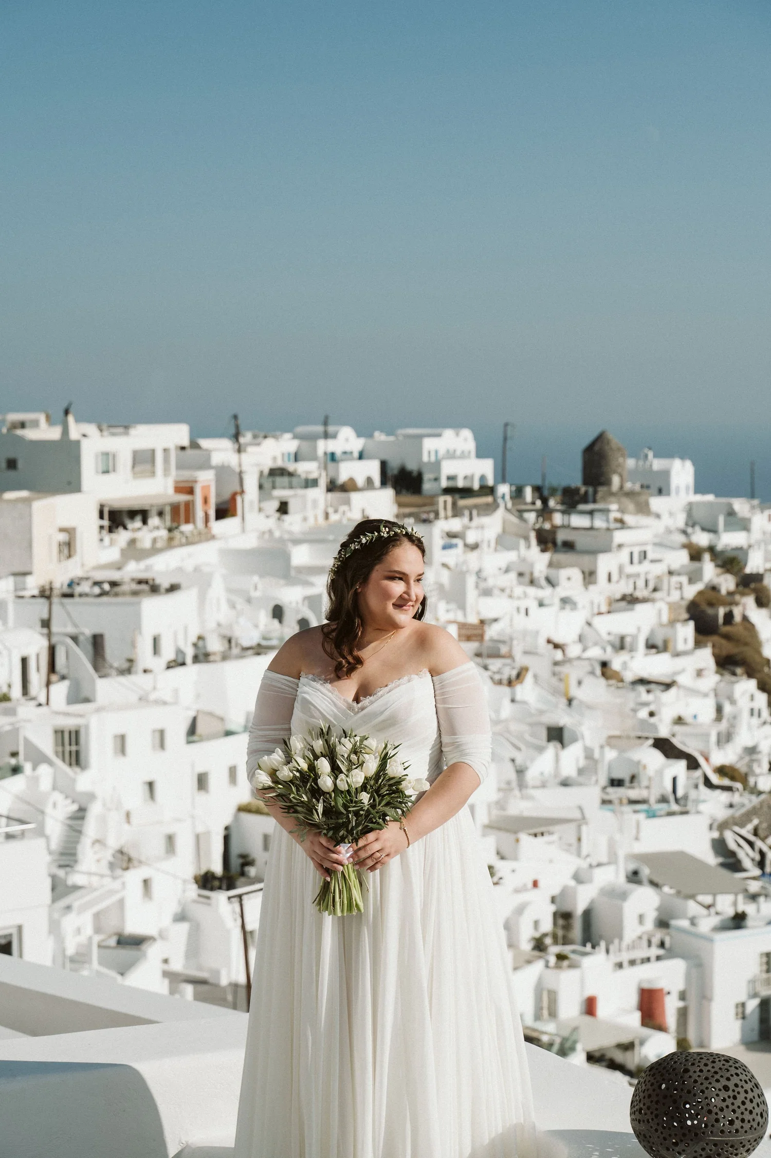 A woman in a wedding dress holding a bouquet of white flowers, standing on a terrace with white buildings and blue sky in the background.