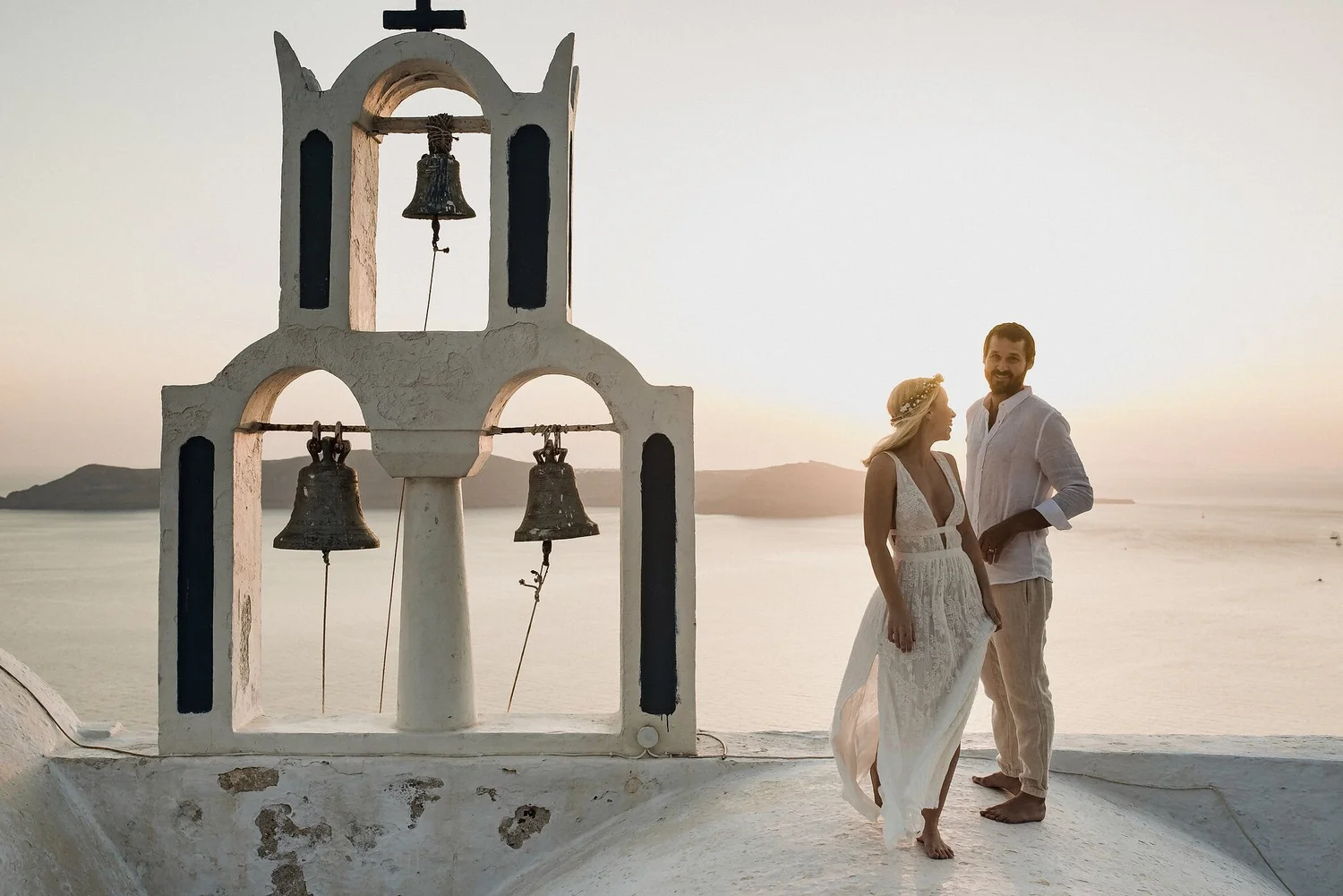 A couple dressed in white wedding attire standing outdoors near a white bell tower with three bells, against a scenic sunset with distant mountains and water in the background.